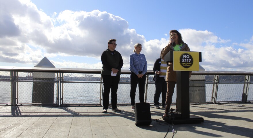 A speaker stands behind a lectern backed by  three other people standing on a pier with water in the background