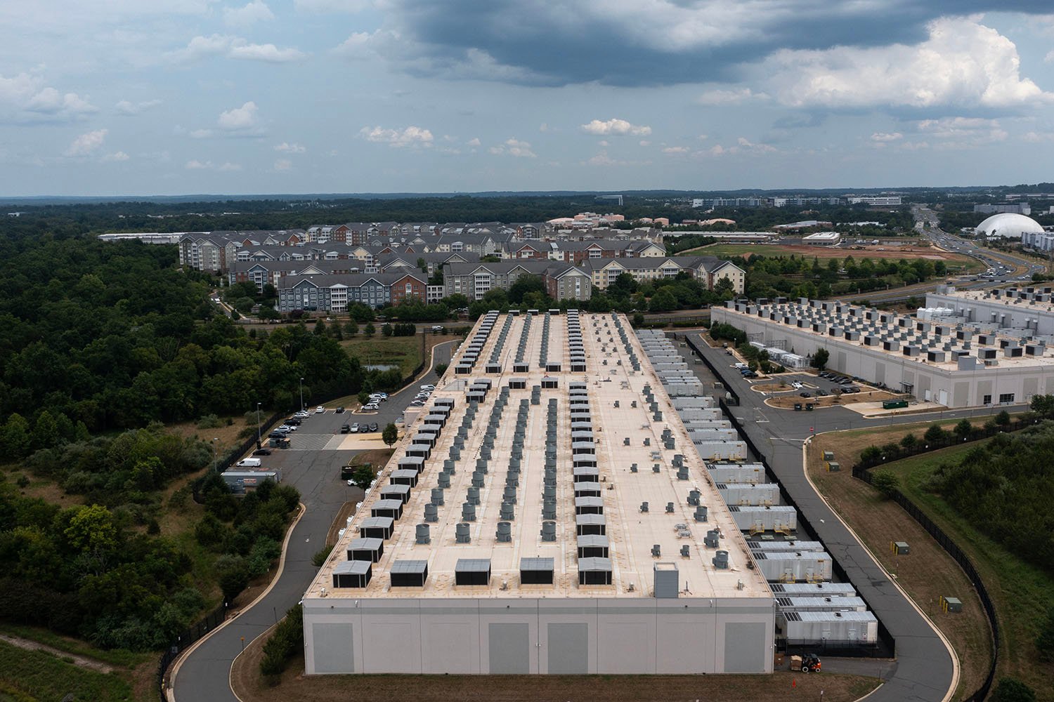 An aerial view shows a giant rectangular building. Suburban houses and trees can be seen in the distance.