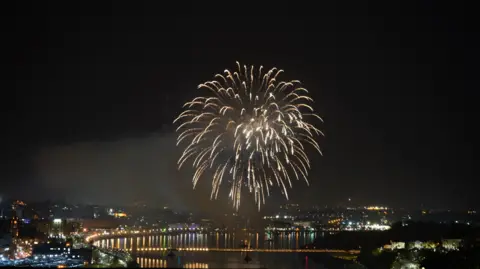 PA Media A singular large firework explodes over the Foyle Bridge in Londonderry. The city is in darkness bar the lights of the bridge and buildings, which are reflected in the River Royle.