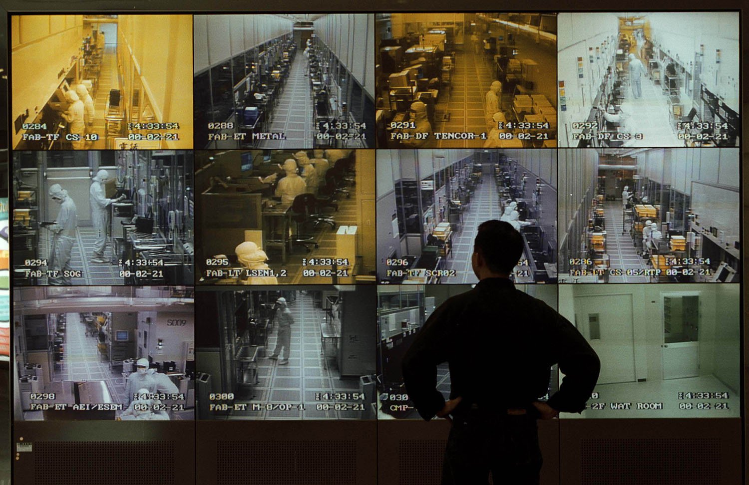 A person with their hands on their hips is silhouetted in front of a bank of monitors showing different views of manufacturing rooms.