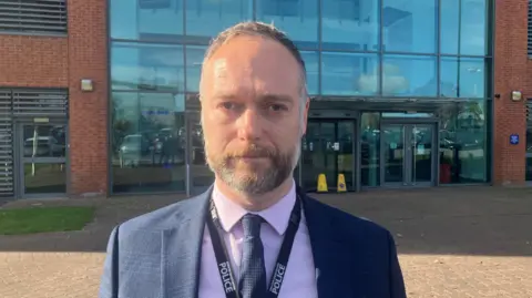  Photo of Detective Superintendent Joe Harrop from GMP outside his office. He is wearing a lilac shirt and navy tie and blazer. He has a greyish beard with short, light hair and is looking into the camera.