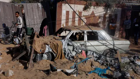 Reuters Palestinians stand near a destroyed car at the site of an Israeli strike on a house in Khan Younis, in the southern Gaza Strip (25 October 2024)