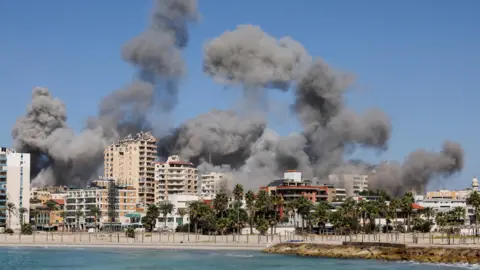 Reuters Smoke billows after Israeli strikes in the southern Lebanese city of Tyre (23 October 2024) - the coast of Tyre is visible from the water, with a number of mid-sized tower blocks and trees visible on Tyre's beach.