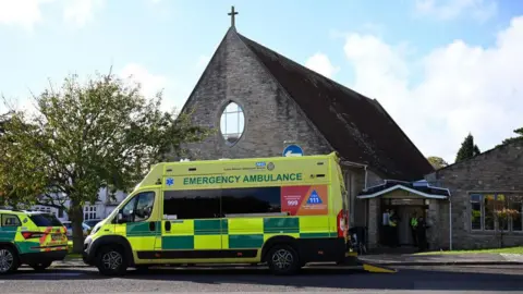 Getty Images An ambulance parked outside All Saints Church in Swanage - a stone-built building with a cross on the roof and an elliptical window on the gable end