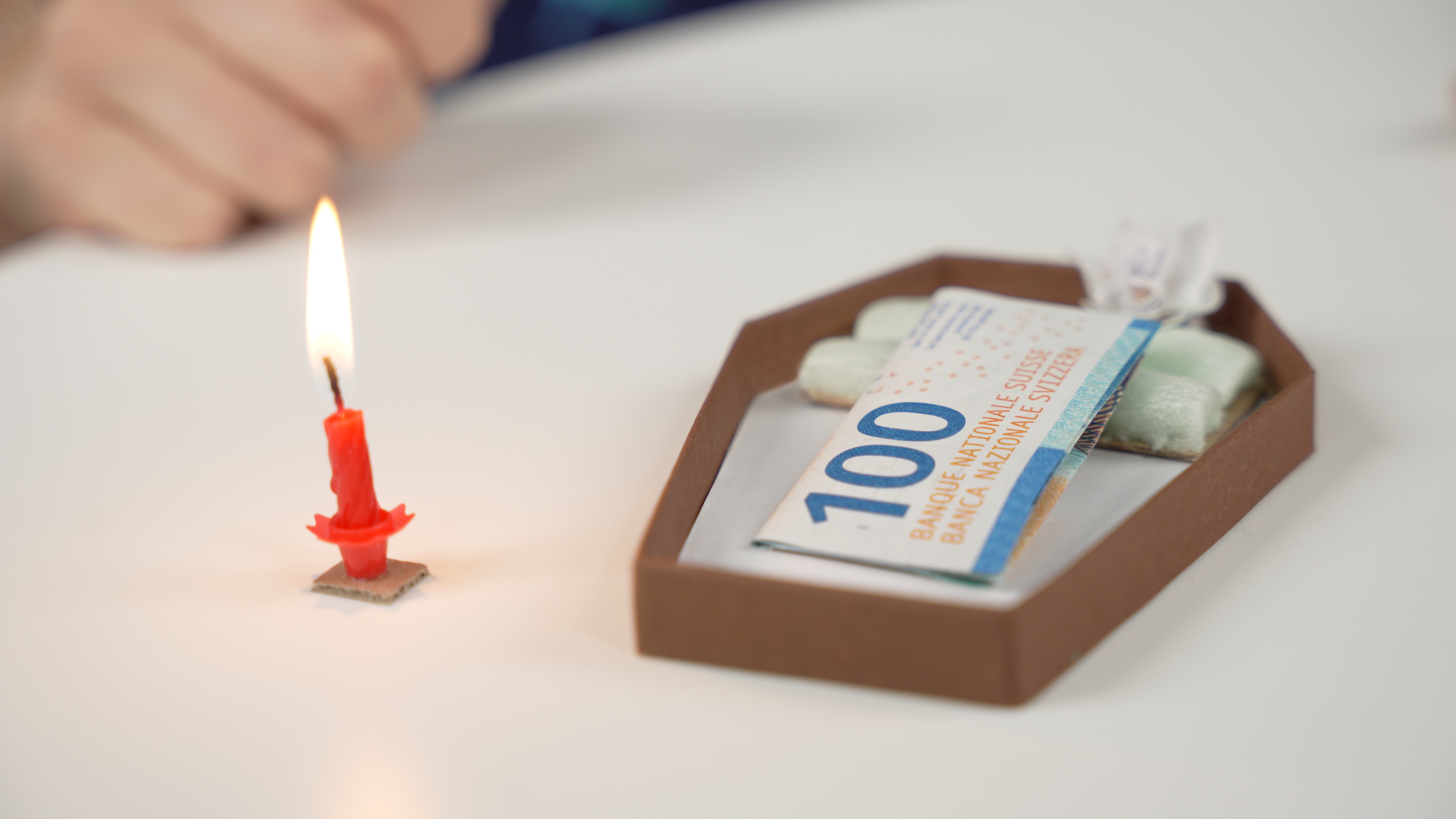 A CHF100 banknote lying in a cardboard coffin next to a lit candle