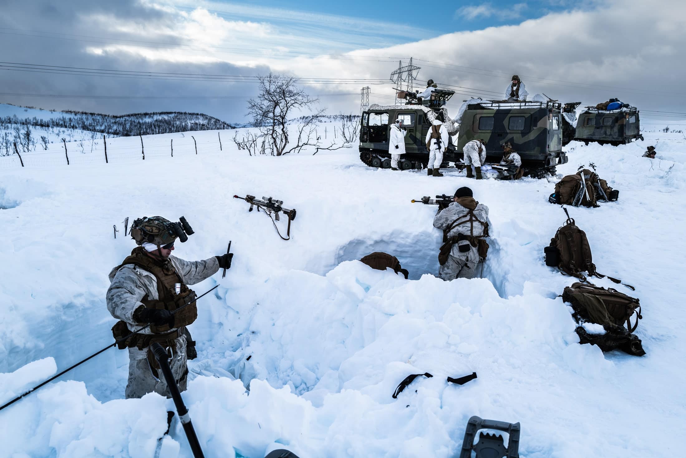 Soldiers in white gear use military equipment in a snowy field with trees and a truck in the background