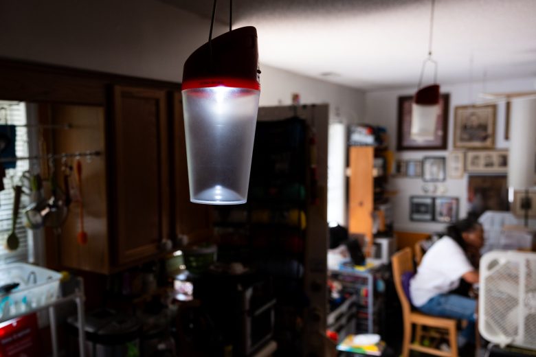 A solar light contained in a plastic cylinder with a red top, hangs in a kitchen. In the background someone is working at a table.