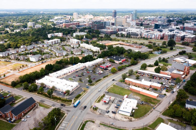 An aerial view of Durham's Hayti Community and beyond. The image shows buildings, highways and parking lots in the foreground. There are large buildings and tree canopy in the background.