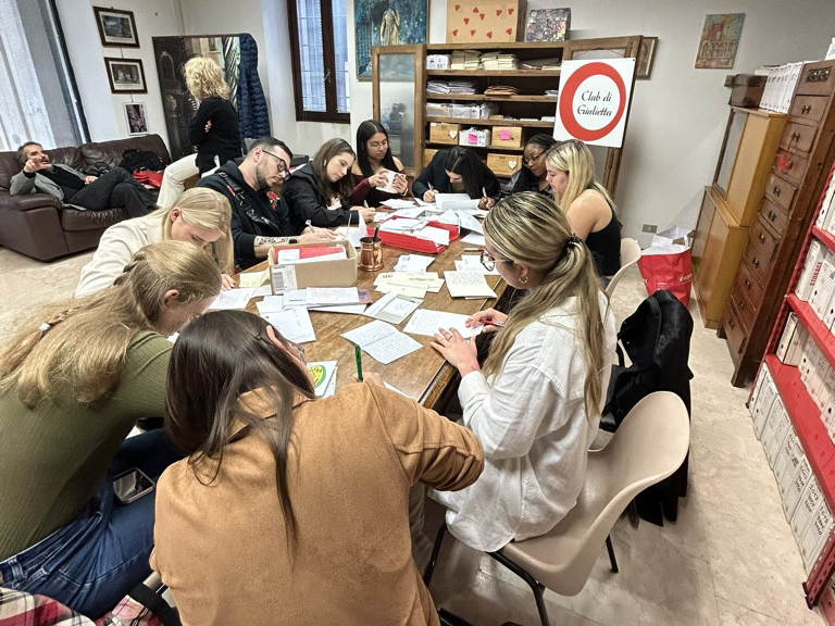 A group of people sit around a table, each person leans forward in concentration over their work as they hand-write letters.