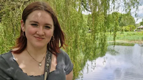 BBC Faye Cullum, wearing a green shirt and a bag strap across her front, stands in front of a lake with tree branches hanging over 