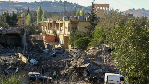 Getty Images People inspect the destruction at the site of an Israeli air strike on the Gouraud Barracks area of Baalbek, with the city's Roman ruins visible in the background (29 October 2024)