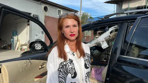 BBC / Imogen Anderson Olena Kryvchun, a woman with red hair and her glasses perched on her head, wearing a white shirt with zebras on it and bright red lipstick, stands by her car. A smashed windscreen, collapsed roof and an interior torn by shrapnel are visible.