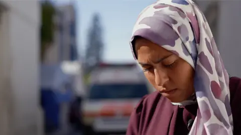 BBC Nevine Al Dawawi wears a lilac and white headscarf and a maroon top. 