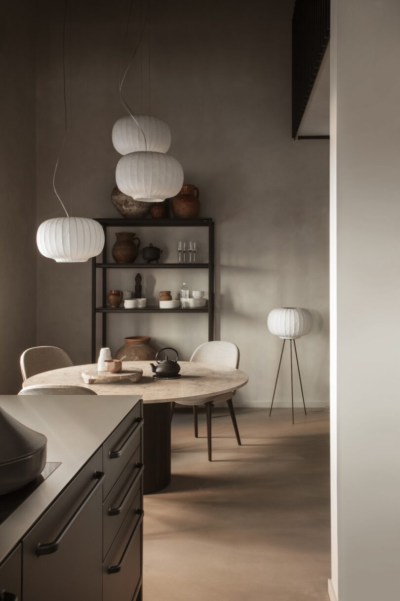 A dining space with three hanging lanterns, a round table, and rustic pottery on a black shelf