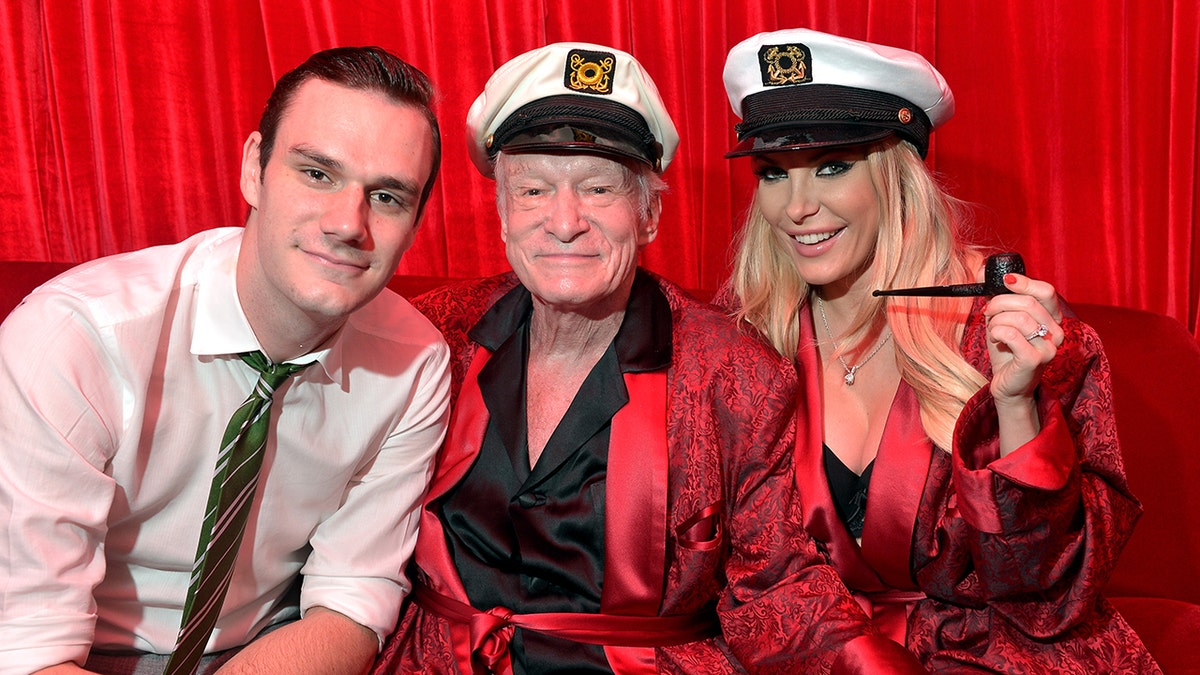 Cooper Hefner, in a white shirt smiles with father Hugh Hefner in a red rob and sailors hat next to wife Crystal Hefner in a similar outfit 