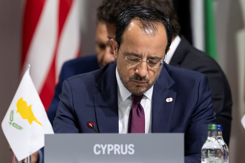 A man in a suit and glasses looking down sitting behind a placard that says cyprus. 