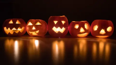 Getty Images A line of pumpkins with different facial carvings, some with eyes and teeth, others with eyes, teeth and a nose - all lit up in front of a black background. The light is reflecting off the floor. 