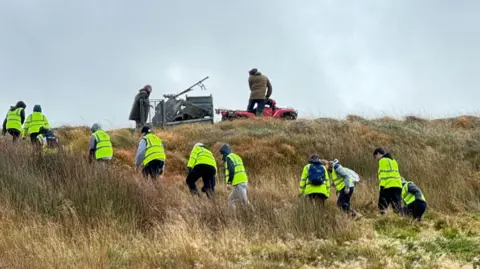 Jonny McNee A line of teenagers in high viz jackets walking up a hill, in the backround are two men, one on a red quead towing the wreck. 