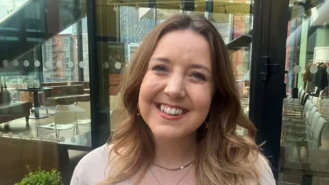 Photo of Amy Bond from Causeway. She has brown curly hair, is wearing red lipstick and a gold necklace and is smiling into the camera. 