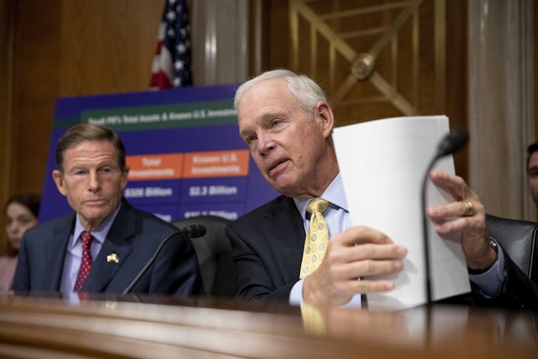 One man in a suit holds up documents during a hearing while another man in a suit looks on.