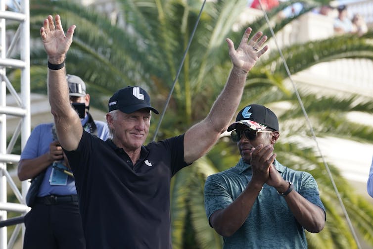 A white man stands with his arms raised triumphantly beside a racialized man. Both wear ballcaps and stand in front of a large palm tree.