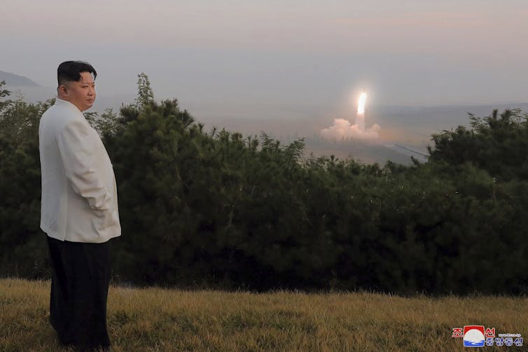 An Asian man in a white jacket stands on a hill overlooking what appears to be a missile launch in the distance.