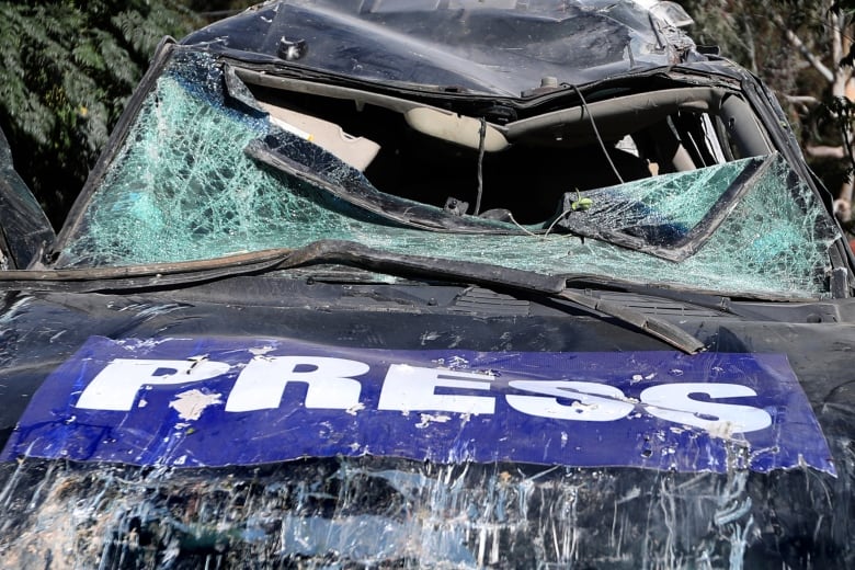 A heavily damaged vehicle with a broken windshield is shown in closeup, with a banner reading 'Press' over its hood.