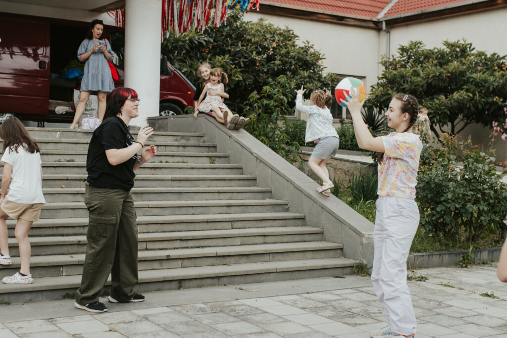 A college student and a girl toss a beach ball to each other outside. Other children are seen playing on the stairs in the background.