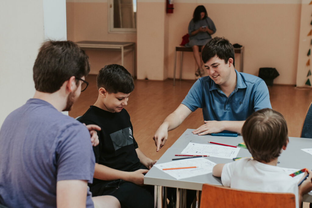 A Mercer student and boy look down while the student points at something unseen in the child's lap. Papers and pencils are on the table in front of them. 