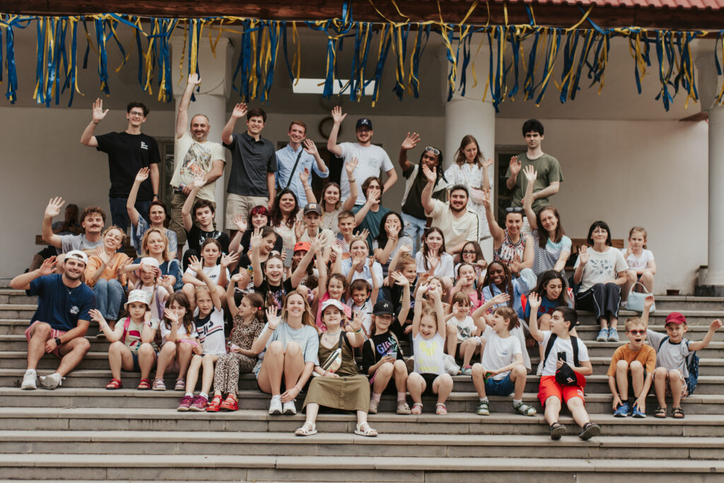 A diverse group of people of various ages sitting and standing on outdoor steps, smiling and waving at the camera.