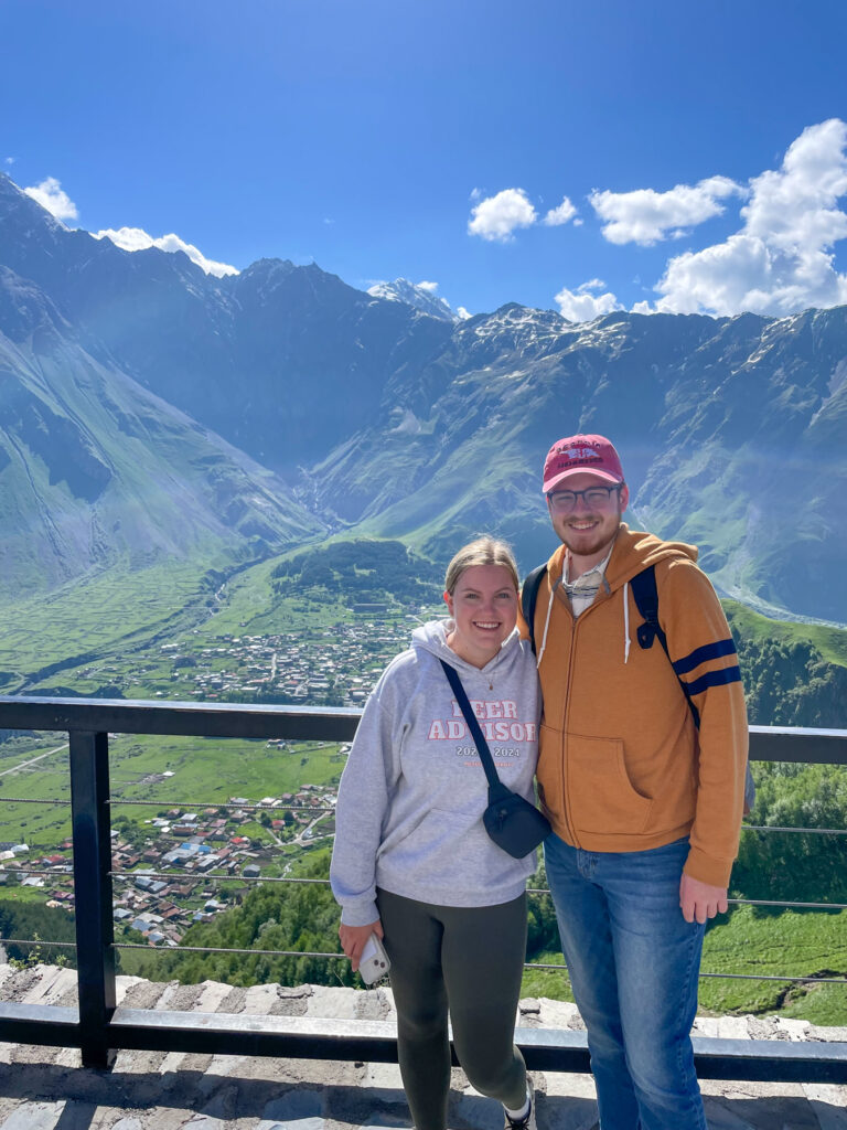 Anna Hale and Harris Wallace wearing casual, long-sleeved shirts and pants in front of a green mountain range and blue sky.