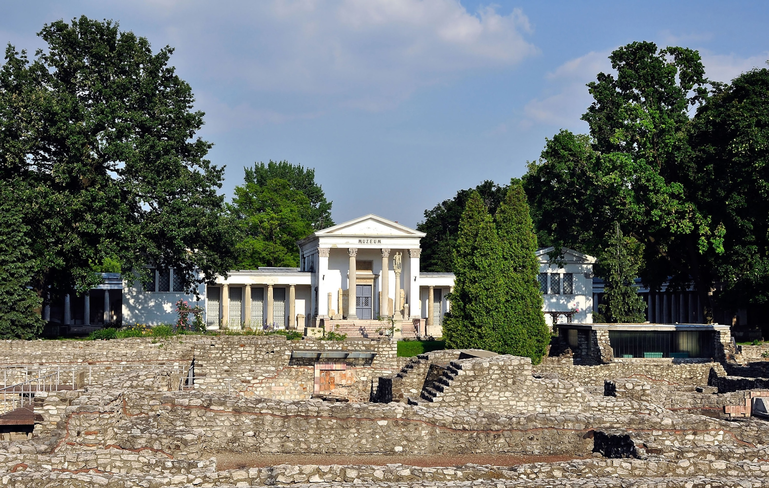 A white museum with columns sits atop a hill. There are ruins in front of it. The sky is blue and there are green trees surrounding the area.