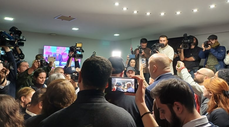 A group of journalists and politicians watch TV and mobile phone screens.