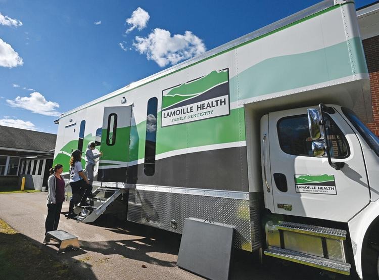 Mobile dental clinic truck with "Lamoille Health Family Dentistry" logo. Two people stand outside, one entering via steps. Sunny day with scattered clouds.